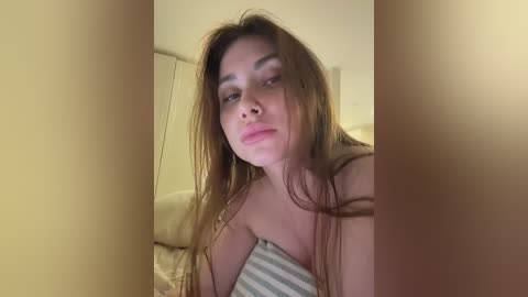A video of a young woman with long, light brown hair, light skin, and a slender build, wearing a striped top. She is indoors, with a blurred background featuring white walls and a bed.