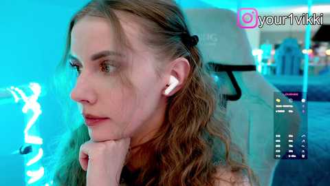 Video of a Caucasian woman with wavy brown hair, wearing white earbuds, deep in thought, sitting in a dimly lit room with blue ambient lighting.