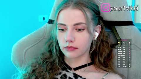 Video of a young, fair-skinned woman with long, wavy brown hair, wearing a black and white patterned top, sitting in a beige gaming chair, with a blue background, and a watermark reading \"@yourvrkitty\" in the top right.