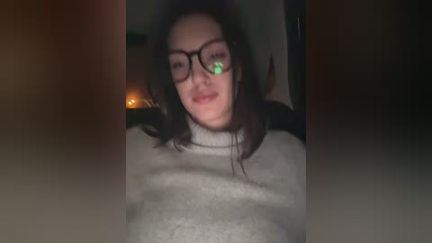 A video of a young woman with straight black hair, wearing glasses and a grey sweater, in a dimly lit room with a lit candle in the background.