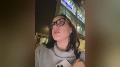 Video of a young woman with dark hair, wearing glasses and a gray sweater, standing in a dimly lit, possibly urban, indoor setting with blurred background lights.