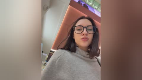 Video of a young woman with straight, dark hair, wearing glasses and a light gray sweater, standing in a modern, well-lit room with a green ceiling and pink accents.