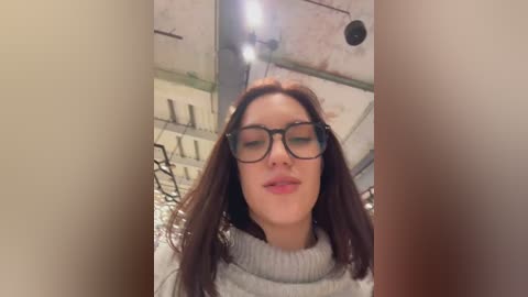 Video of a young woman with long brown hair, wearing black-rimmed glasses and a light grey sweater, standing in a dimly lit, industrial-style room with exposed pipes and metal shelves in the background.