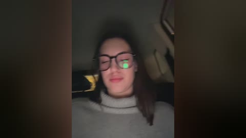 A blurry video of a woman with straight brown hair, wearing glasses and a grey sweater, seated in a dimly lit car with a green light reflecting on her face.