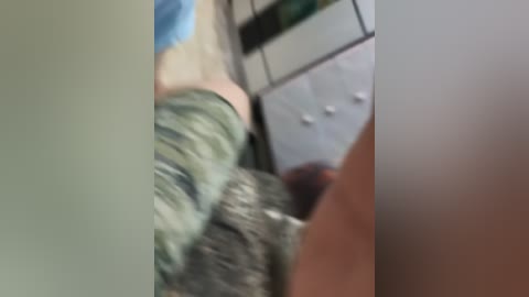 A blurry video of a person wearing green camo pants and a blue shirt, standing near a white dresser with silver handles in a dimly lit room.