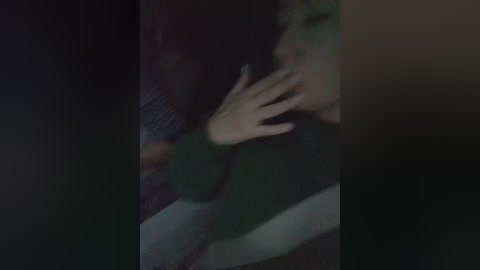 A dimly-lit video showing a person with light skin and short, dark hair, wearing a dark sweater, with their hand over their mouth, in a dimly-lit room.