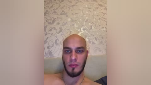 Video of a bald, light-skinned man with a trimmed beard, wearing a necklace, seated against a beige wall with a floral-patterned wallpaper.