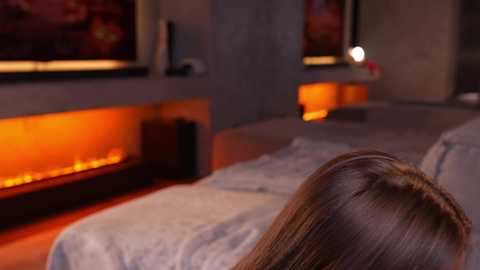Video of a dimly lit bedroom with a warm, orange-tinted fireplace on the wall, featuring a brunette woman lying on a bed with gray bedding, partially visible.
