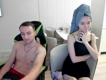 Video of a shirtless man with short brown hair and a woman with blue towel-wrapped hair, both seated in modern chairs in a minimalist room with white walls and vertical blinds.