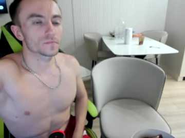Video of a shirtless, muscular man with short hair, wearing a silver necklace, sitting on a green gaming chair. Background includes a modern dining area with white chairs, a white table, and a glass of water.