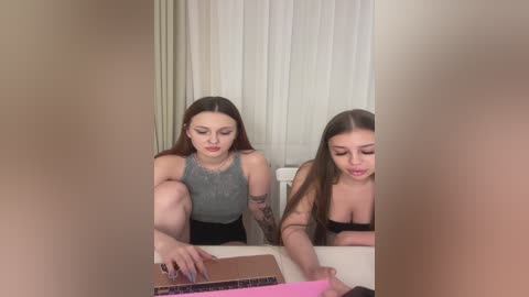 A video showing two young women with long hair, tattoos, and wearing casual outfits seated at a table, with a blurred figure in the foreground.