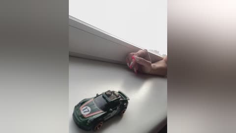 A video showing a person's hand with painted nails reaching through a window frame, holding a green toy car with the number 11 on it. The background is bright, with a sunlit room and soft lighting.