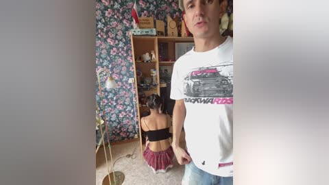 Video of a young woman with dark hair, wearing a black bra and red skirt, kneeling on a carpet in a room with floral wallpaper and wooden shelves, a man in a white t-shirt stands nearby.