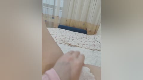 Video of a person in a bed with white sheets, holding a pink sock, against a backdrop of a window with sheer curtains.