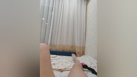 A video shows a woman lying on a bed with white floral sheets, her legs spread, and a black hairband near her feet. The room has white walls, a sheer white curtain, and a blue headboard.