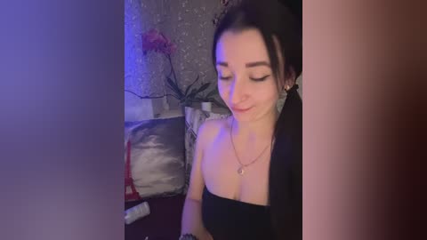 Video of a smiling, fair-skinned woman with dark hair, wearing a black strapless top, seated on a gray couch with a red bag and a potted plant nearby.