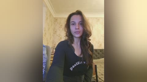 Video of a young woman with long, wavy brown hair, wearing a black \"Pacific Northwest\" shirt, standing in a bedroom with floral wallpaper and a bed.