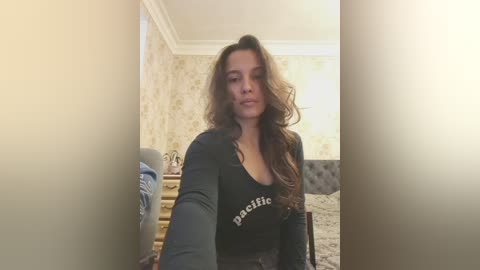 Video of a brunette woman in a black top, standing in a beige-patterned room with a bed, lamp, and staircase in the background.