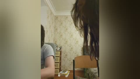 A video depicting a woman with long brown hair, seated at a wooden table in a dimly lit room with floral wallpaper and a television.