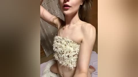 A video of a slender, fair-skinned woman with long brown hair, wearing a white, fuzzy, strapless bandeau top and a delicate necklace. She is indoors, partially obscured by a beige curtain.