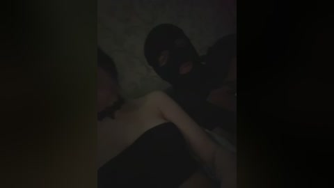 A dimly lit video of a person wearing a black mask, lying on a bed with a floral-patterned bedspread. The figure is partially obscured, suggesting an intimate or secretive atmosphere.