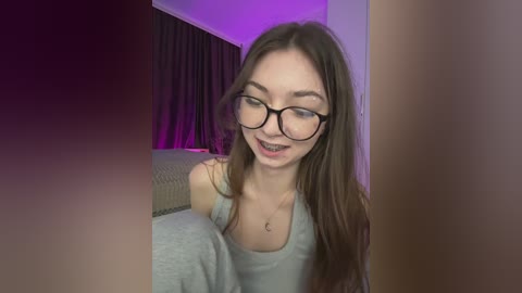 Video of a young, slender woman with long brown hair, wearing glasses and a gray tank top, smiling, in a dimly lit bedroom with purple lighting.