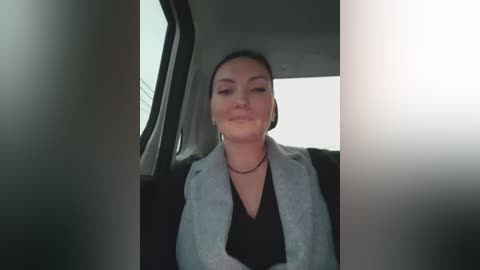 Video of a woman with light skin and dark hair, wearing a black top and light gray blazer, sitting in a car.