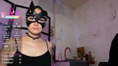 Video of a young woman with light skin, wearing a black cat mask, black top, and choker, in a dimly lit room with a sink and cluttered countertop.