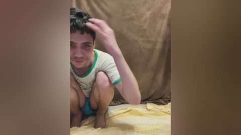 Video of a young boy with short dark hair, wearing a white T-shirt and blue shorts, squatting on a yellow bedspread, with a beige fabric backdrop.