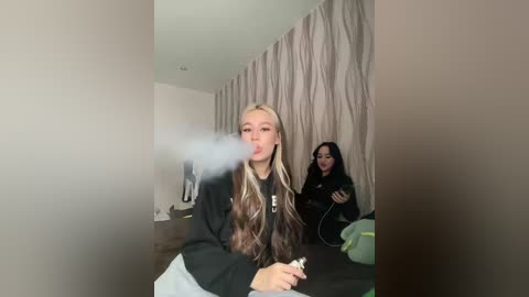 Video of a blonde woman in a black hoodie blowing smoke, indoors, with another person in the background.