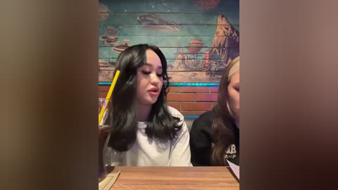 Video of a young woman with long, dark hair, wearing a white hoodie, sitting in a booth with a yellow pencil in her mouth. Background features a mural with underwater scenes.