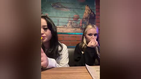 Video of two young women with long hair, one brunette and one blonde, sitting at a wooden table, eating with their hands, in a dimly lit restaurant with a mural of a beach and mountains on the wall.