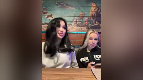 Video of two young women with long, wavy hair, one with dark hair and the other with blonde streaks, both wearing black T-shirts with white lettering, sitting at a wooden table in a dimly-lit, nautical-themed bar.