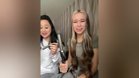 Video of a young woman with long blonde hair, wearing a green shirt, holding a hairdryer, standing next to a young Asian woman with long black hair, dressed in a white shirt, both smiling.