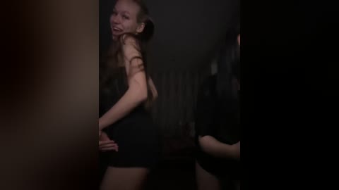 Video of a slender, light-skinned woman with long, straight brown hair, wearing a black, sleeveless dress, standing in a dimly lit, shadowy room. Her expression is playful.