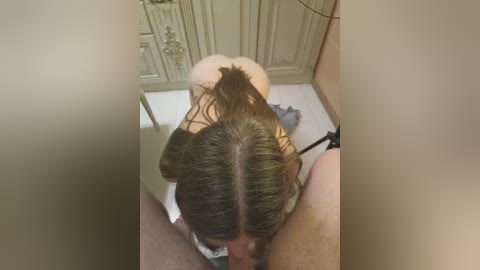Video of a woman with long, wet hair, kneeling on a bathroom floor, performing oral sex on a man; beige cabinets in the background.