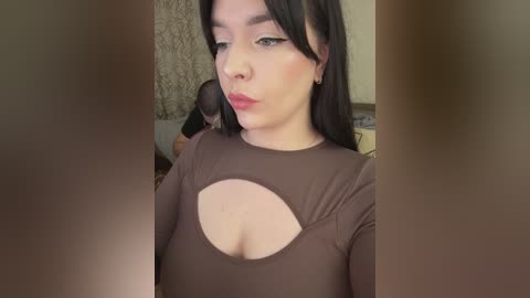 Video of a light-skinned woman with long dark hair, wearing a brown, long-sleeved top with a large cutout revealing her cleavage. Background shows a textured wall and a blurred figure.