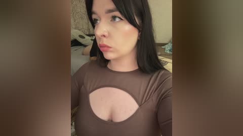 Video of a light-skinned woman with long black hair, wearing a brown top with a large cutout revealing cleavage, in a dimly lit bedroom.