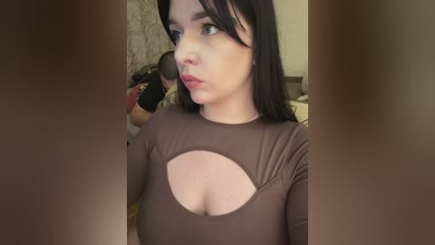 A video of a young woman with pale skin, black hair, and a brown top with a large cut-out. She looks contemplative. Background includes a messy room with a bed and a person partially visible.