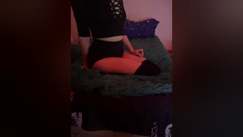 Video of a woman with fair skin, wearing a black crop top and high-waisted black shorts, kneeling on a green bedspread in a dimly lit room.