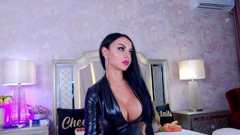 Video of a Latina woman with long black hair, wearing a black, low-cut leather dress, standing in a stylish bedroom with a silver headboard, two bedside lamps, and decorative pillows.