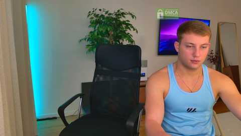 Video of a fit young man in a blue tank top, sitting in a black mesh chair in a modern living room with a green potted plant, TV, and wall-mounted art.