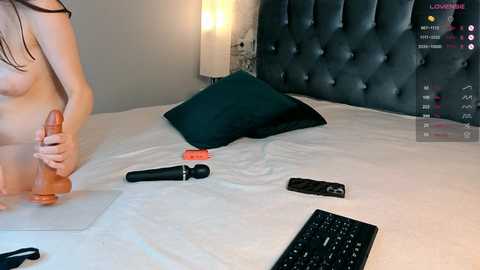 A video of a nude woman with long brown hair, holding an orange dildo, on a bed with a black tufted headboard. A black keyboard and remote controls are on the bed, with a black dildo and a red vibrator nearby.