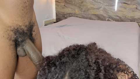 A video showing a close-up of a dark-skinned man's erect penis with a hairy pubic area, positioned over a light-skinned woman's dark curly hair, both on a white bed in a room with beige stone walls.