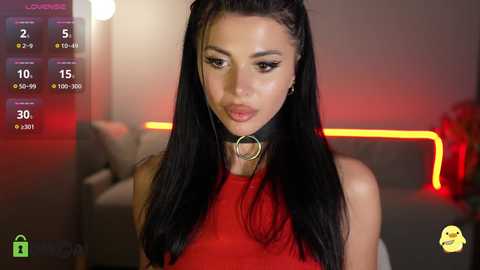 A video of a woman with long black hair, wearing a red top and black choker, in a dimly-lit room with neon lighting.
