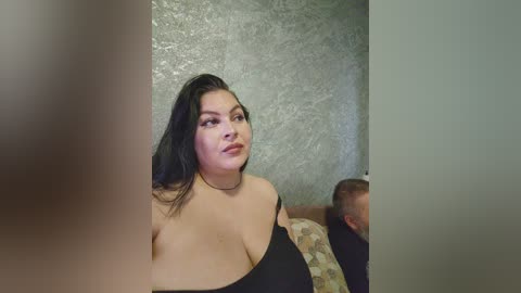 Video of a curvy woman with long black hair, wearing a black off-shoulder top, sitting on a patterned couch, against a textured wall background.