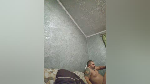 Video of a man with a shaved head, muscular build, and tattoos, lying on a patterned couch in a dimly lit room with textured grey walls and a ceiling.
