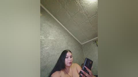 Video of a young woman with long black hair, medium build, and tan skin, taking a selfie in a dimly lit room with textured, light gray walls and a white ceiling. She wears a black top.