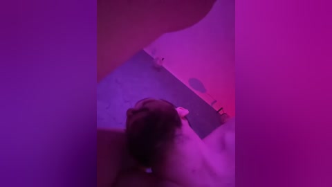 A video featuring a nude woman with dark hair, lying on a bed, partially obscured by neon purple and pink lights. The image is slightly out of focus, creating a dreamy, surreal atmosphere.