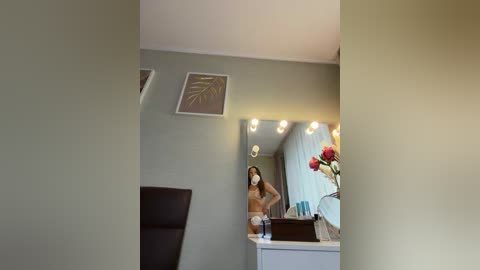 Video of a modern bathroom with beige walls, a mirror with multiple lights, a white vanity with a red vase, a black chair, and a framed leaf design on the wall.